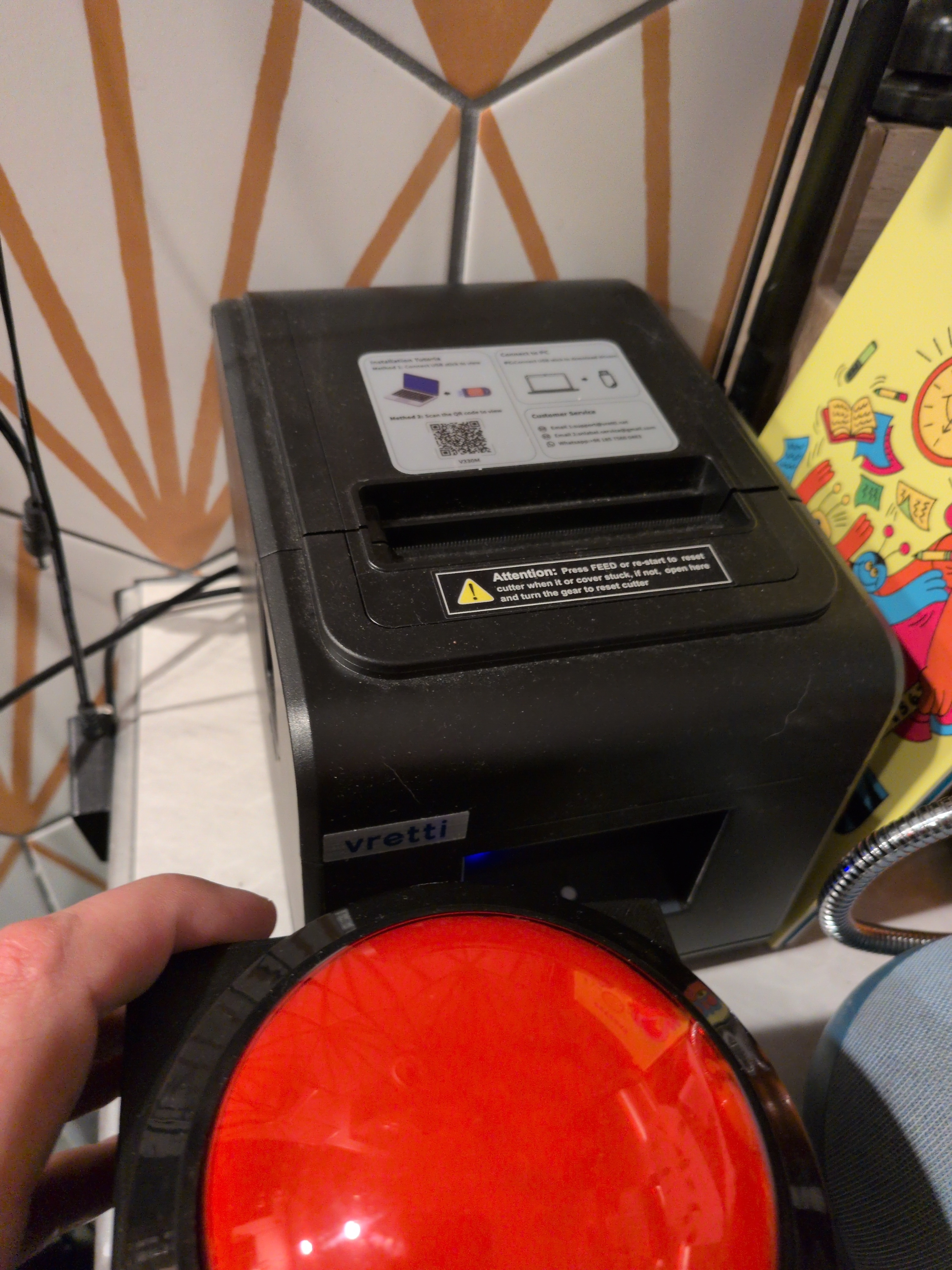 Red button on top of a black box with a black receipt printer  behind it