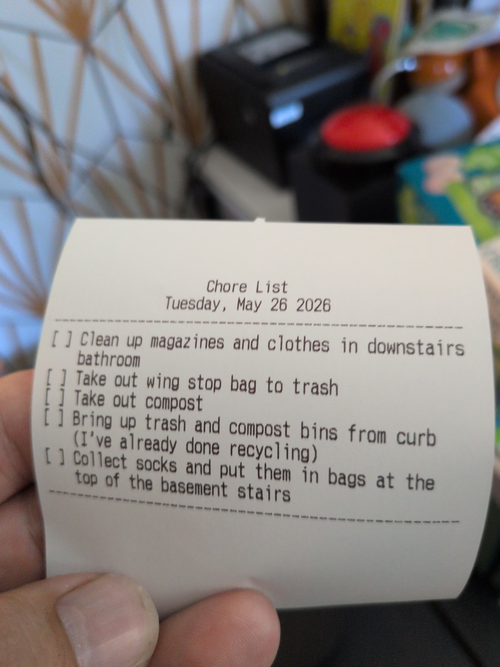 A paper receipt with the title 'Chore List' 'Tuesday, May 26 2026' and the chores: 'Clean up magazines and clothes in downstairs bathroom', 'Take out wing stop bag to trash', 'Take out compost', 'Bring up trash and compost bins from curb', and 'Collect socks and put them in bags at the top of the basement stairs'